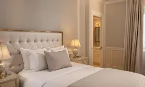 Elegancka sypialnia z dużym łóżkiem, pikowanym zagłówkiem i stylowymi lampkami nocnymi. Widok na łazienkę i zasłony. Idealne miejsce dla wynajmującego szukającego komfortu.