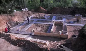 Budowa fundamentów domu. Widać zbrojenie i szalunki. W tle koparka i materiały budowlane, w tym chudy beton.
