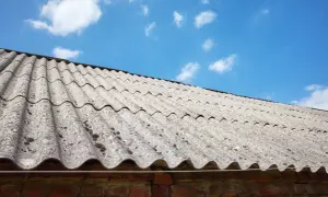 Stary dach pokryty eternitem, z widocznymi śladami mchu, pod błękitnym niebem z chmurami.