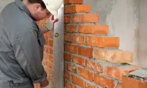 Murarz w kasku i rękawicach sprawdza pion muru za pomocą poziomicy. Widać kolejne warstwy cegieł, których wymiary są kluczowe dla stabilności konstrukcji.