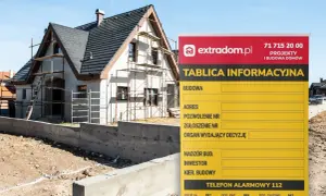 Tablica informacyjna budowy domu w trakcie budowy, z widocznym rusztowaniem i fundamentami. Na tablicy dane inwestora i numer telefonu alarmowego.