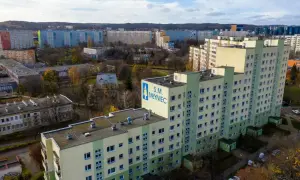 Osiedle z wielkiej płyty S.M. Młyniec, z charakterystycznymi blokami w kolorach zieleni i beżu, otoczone drzewami w jesiennej szacie.