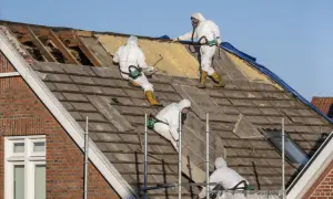 Trzech pracowników w kombinezonach ochronnych usuwa stare pokrycie dachowe, które może zawierać szkodliwy **azbest**.