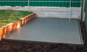Świeżo wylana płyta betonowa z widocznym zbrojeniem rozproszonym, otoczona drewnianą palisadą i zielonym ogrodzeniem.