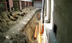 Prace przy odwodnieniu domu. Wykopano rów, w którym ułożono nowe, pomarańczowe rury. Obok leżą stare, zniszczone rury.