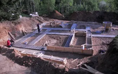 Budowa fundamentów domu. Widać zbrojenie i szalunki. W tle koparka i materiały budowlane, w tym chudy beton.