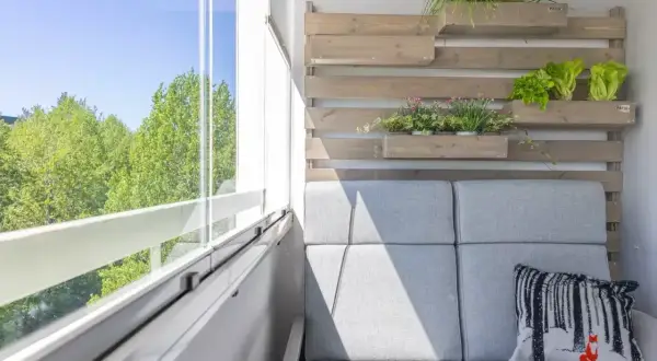 Przytulna loggia z szarą sofą, poduszką z liskiem i zielenią na drewnianej ścianie. Widok na drzewa przez przeszklony balkon.