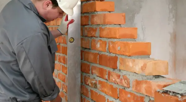 Murarz w kasku i rękawicach sprawdza pion muru za pomocą poziomicy. Widać kolejne warstwy cegieł, których wymiary są kluczowe dla stabilności konstrukcji.