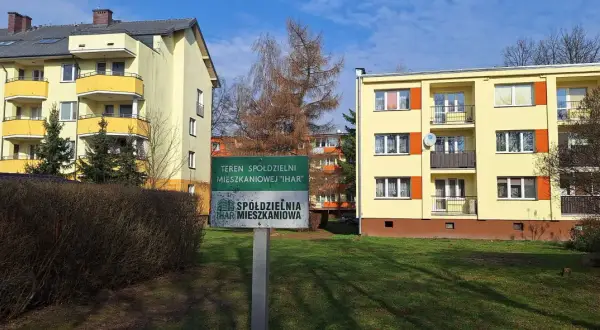 Zielony znak z napisem "TEREN SPÓŁDZIELNI MIESZKANIOWEJ 'IHAR'" przed żółtymi blokami mieszkalnymi.