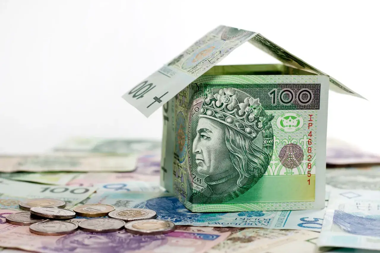 Dom z banknotów 100 zł, symbolizujący mieszkanie na start. Na pierwszym planie monety.