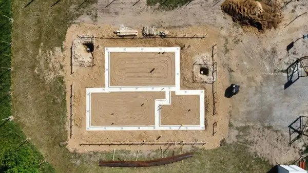 Widok z lotu ptaka na budowę domu. Widać gotowy fundament, piasek, wykopy i materiały budowlane.