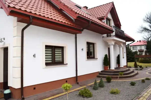 Biały dom z brązowym dachem i rynnami. Widoczny cokół na elewacji, ozdobne schody i zadbany ogród.