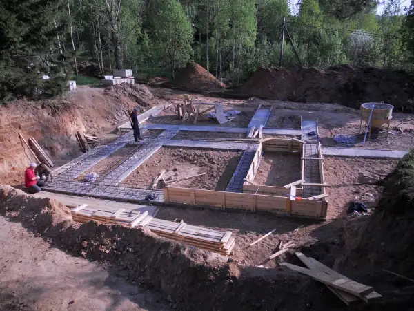 Budowa fundamentów domu. Widać zbrojenie i szalunki. W tle koparka i materiały budowlane, w tym chudy beton.
