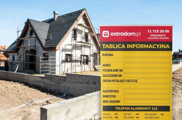 Tablica informacyjna budowy domu w trakcie budowy, z widocznym rusztowaniem i fundamentami. Na tablicy dane inwestora i numer telefonu alarmowego.