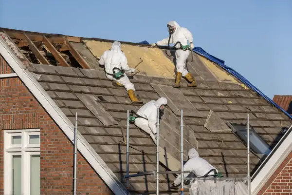 Trzech pracowników w kombinezonach ochronnych usuwa stare pokrycie dachowe, które może zawierać szkodliwy **azbest**.